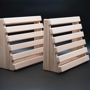 May include: Two wooden display stands. Each stand is constructed from light-colored wood and features a slatted design with horizontal wooden bars. The stands are angled, creating a tiered effect, and are suitable for displaying items.