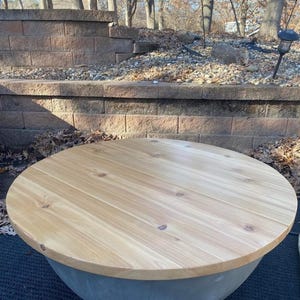 May include: A round fire pit table featuring a smooth, light-toned natural wood top with visible grain and knots, set upon a solid, bowl-shaped grey concrete base. The circular wooden surface is polished and sits flush atop the concrete structure.