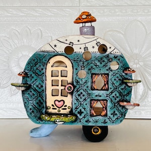 Op de afbeelding: Een grillige keramische camper in tinten van teal, wit en oranje. De camper heeft een gedetailleerde deur, ramen met gordijnen en decoratieve paddenstoelaccenten. Het heeft een paddenstoel schoorsteen en lichtslingers.