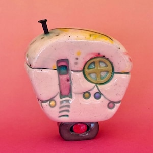 Può includere: Un'eccentrica scatola portagioie in ceramica a forma di casa. La scatola è principalmente bianca con accenti colorati, tra cui una porta rosa, una finestra verde e oro e piccoli punti colorati. Un dettaglio nero a forma di antenna si trova sulla parte superiore del tetto. La base è grigia con un bottone rosso.
