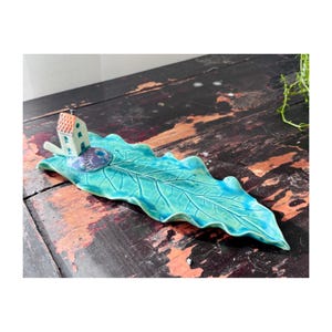 May include: A ceramic leaf-shaped dish in turquoise and teal, with a small house and a blue object on top. The dish has detailed veining and a glossy finish, ideal for decorative use.