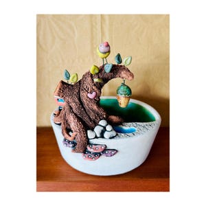Puede incluir: Una maceta de cerámica con forma de árbol con un pájaro posado en una rama. La maceta tiene una abertura en forma de corazón y una pequeña bellota verde colgando de una rama. La maceta está pintada en tonos de marrón, verde y azul.
