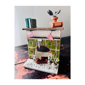 May include: A miniature ceramic fireplace with a green tiled facade and a wooden mantel. The mantel holds small books, a potted plant, and pink triangle banners. The fireplace has a detailed interior with a log and a small plant.