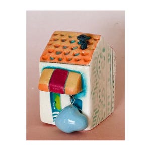 Op de afbeelding: Een klein keramisch huis met een rood en geel luifel, een blauwe bel die aan het dak hangt en een witte en groene gepatroneerde gevel.