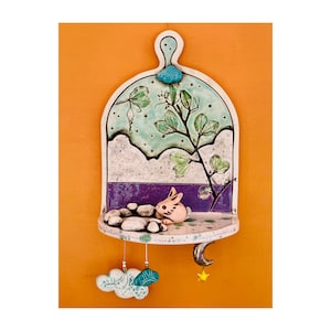 May include: A ceramic wall shelf with a whimsical design featuring a green plant, a pink bunny, and a crescent moon. The shelf has a light blue and green background with white clouds and stars.