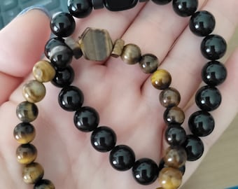 Puede incluir: Dos pulseras, una negra con un dije cuadrado y otra con cuentas de ojo de tigre marrón y dorado.