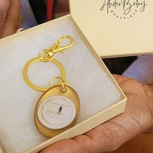 May include: A gold-toned keychain with a circular pendant featuring a white center insert printed with the text Coach & Mentor Tyesha Bolden. The keychain is presented in a cream-colored box with the Ander Berry Bracelets logo on the lid.