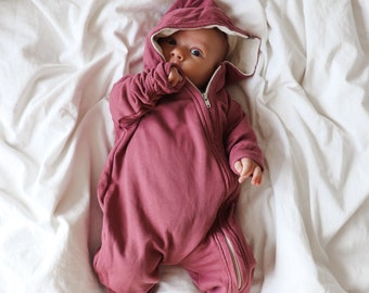 Peut inclure: Un bébé portant une gigoteuse à capuche rose avec une fermeture éclair. Le bébé est allongé sur un drap blanc.