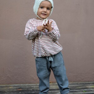 May include: A child wearing a cream-colored knit bonnet, a striped long-sleeve shirt with red stripes, and gray pants with pockets. The child is also wearing rose gold sandals. The background is a solid brown wall.