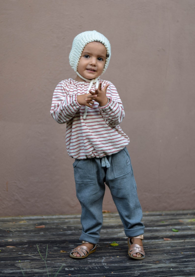 May include: A child wearing a cream-colored knit bonnet, a striped long-sleeve shirt with red stripes, and gray pants with pockets. The child is also wearing rose gold sandals. The background is a solid brown wall.