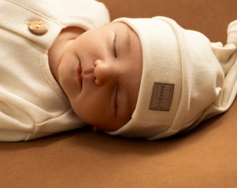 May include: A sleeping baby wearing a cream-colored outfit and a knotted hat. The outfit has wooden buttons. The hat has a small brown label with the text "boxx." The baby is lying on a brown surface.