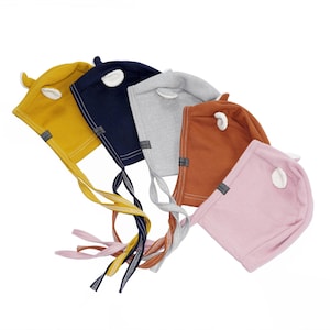 Może przedstawiać: Pięć czapek dla niemowląt w różnych kolorach: żółtym, granatowym, szarym, pomarańczowym i różowym. Każda czapka ma małe białe filcowe ucho na górze i długi sznurek.