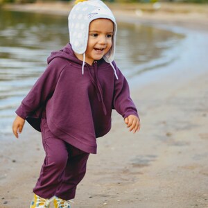 May include: A child wearing a purple hooded poncho and matching pants, paired with yellow and blue sneakers, is running on a sandy beach. The child is also wearing a light blue hat with white polka dots and a yellow pom-pom.