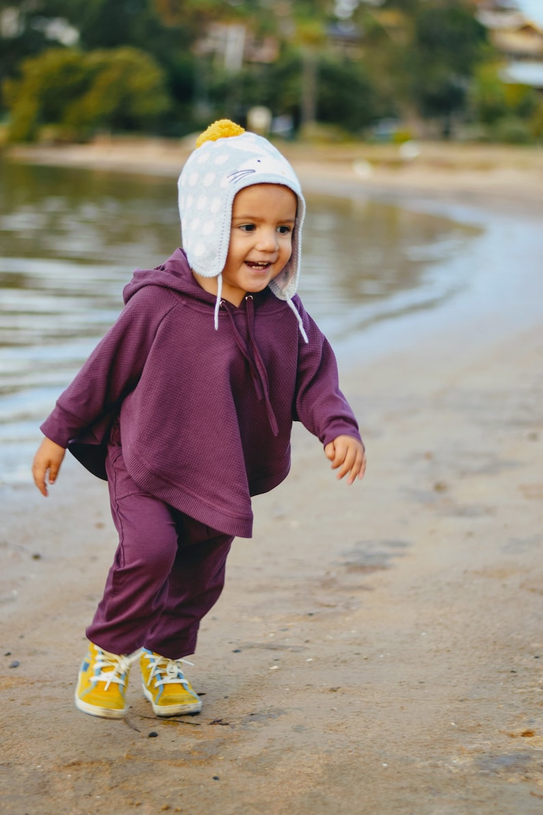 May include: A child wearing a purple hooded poncho and matching pants, paired with yellow and blue sneakers, is running on a sandy beach. The child is also wearing a light blue hat with white polka dots and a yellow pom-pom.