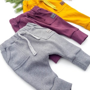 May include: Three pairs of baby pants in mustard yellow, deep purple, and gray. Each pair has a textured fabric, a front pocket, and a drawstring. The pants are displayed on a white surface with evergreen branches.