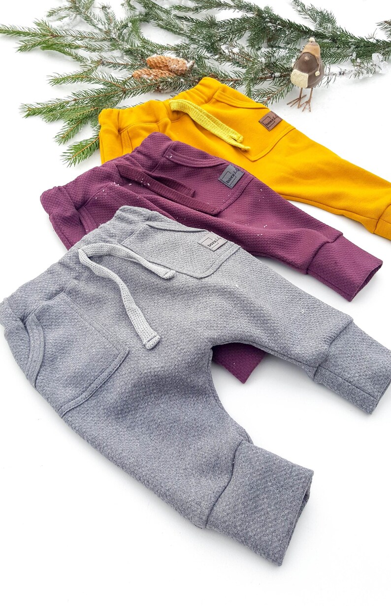 May include: Three pairs of baby pants in mustard yellow, deep purple, and gray. Each pair has a textured fabric, a front pocket, and a drawstring. The pants are displayed on a white surface with evergreen branches.