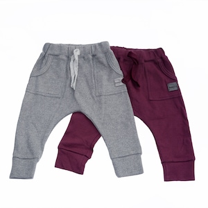 May include: Two pairs of baby pants in gray and burgundy. The gray pants have a drawstring and front pockets. The burgundy pants also have front pockets and a drawstring. Both pairs have a small label on the pocket.