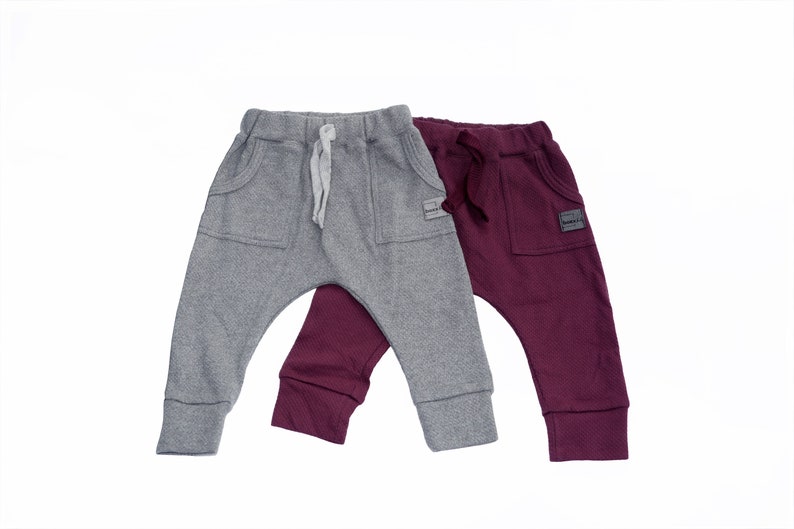 May include: Two pairs of baby pants in gray and burgundy. The gray pants have a drawstring and front pockets. The burgundy pants also have front pockets and a drawstring. Both pairs have a small label on the pocket.