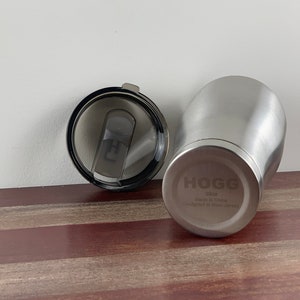 May include: A stainless steel travel mug with a black lid. The mug has the word "HOGG" engraved on the side, along with the text "atlos Made in China Designed in New Jersey".