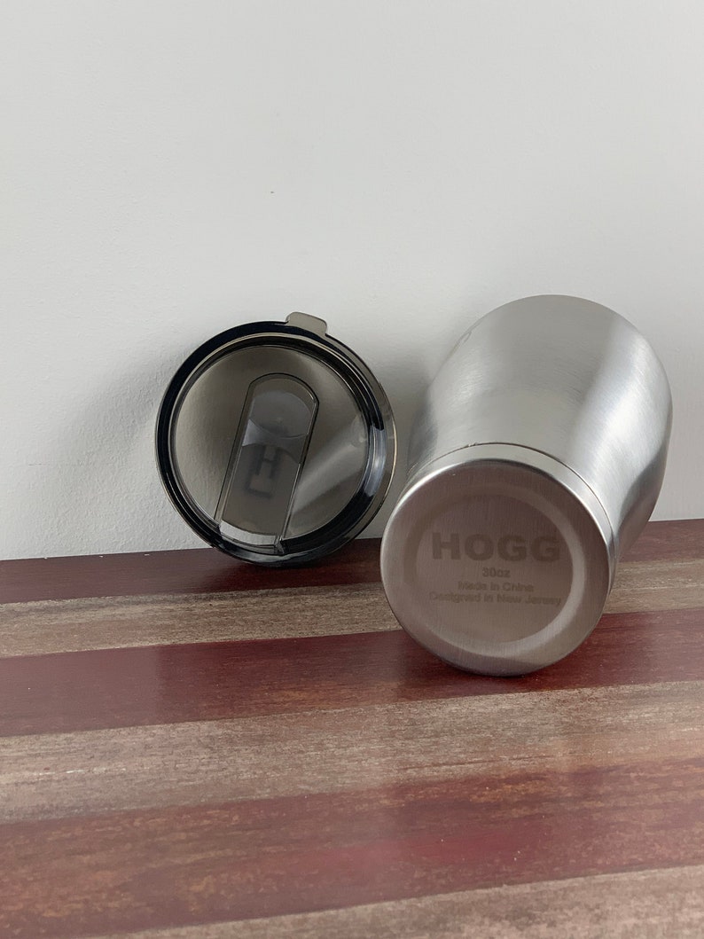 May include: A stainless steel travel mug with a black lid. The mug has the word "HOGG" engraved on the side, along with the text "atlos Made in China Designed in New Jersey".
