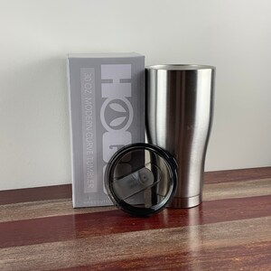 May include: A silver 30 oz. modern curve tumbler with a black lid. The tumbler is sitting on a wooden surface next to a box that says "Hogg Outfitters" and "30 oz. Modern Curve Tumbler".