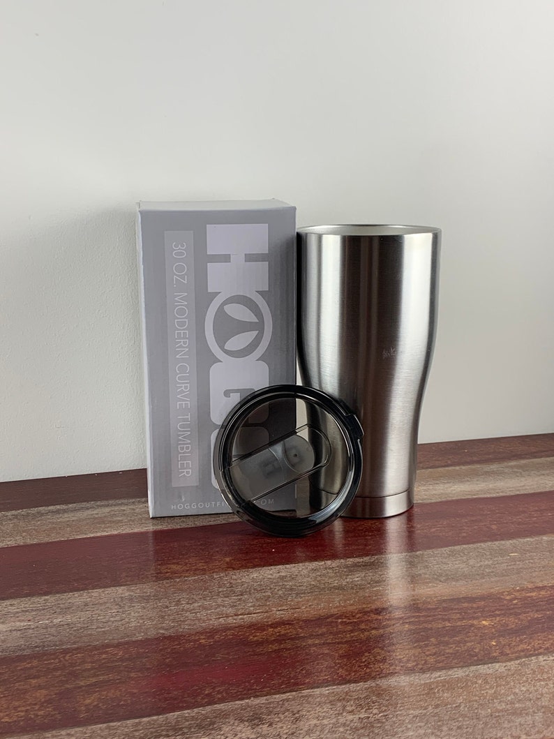 May include: A silver 30 oz. modern curve tumbler with a black lid. The tumbler is sitting on a wooden surface next to a box that says "Hogg Outfitters" and "30 oz. Modern Curve Tumbler".