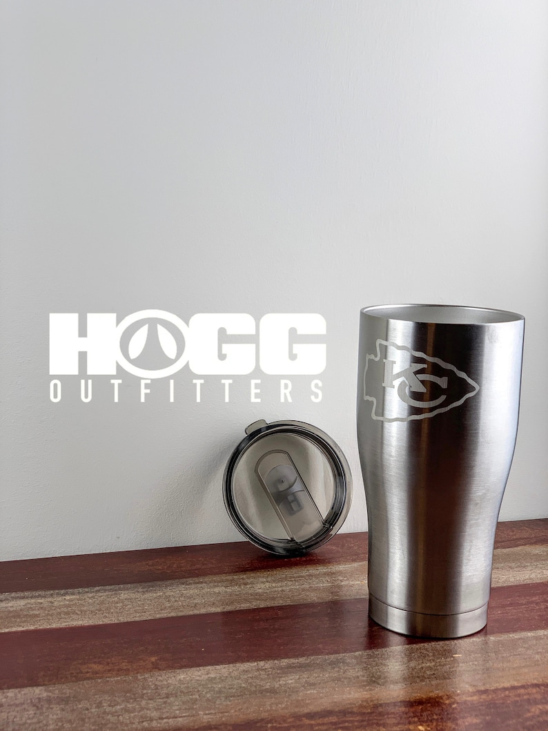 May include: A stainless steel travel mug with a white Kansas City Chiefs logo engraved on the side. The mug is sitting on a wooden surface with a matching lid next to it. The text "HOGG OUTFITTERS" is visible in the background.