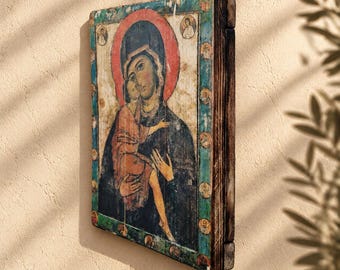 May include: A rectangular wooden icon featuring a painted depiction of a figure holding a child. The central image is framed by a teal border containing small circular portraits. The wood has a dark, weathered finish on the side edges.