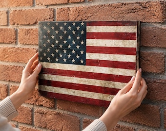 May include: A rectangular wooden sign depicting the American flag. The flag design features red and white stripes and a blue field with white stars, with a distressed, vintage look. The sign is held against a brick wall.