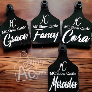 May include: Four black plastic livestock ear tags with a rounded shield shape and a top mounting post. Each tag features the white printed text MC Show Cattle, a logo, and individual names: Grace, Fancy, Cora, and Mercedes. A watermark reads Derholt Customs.