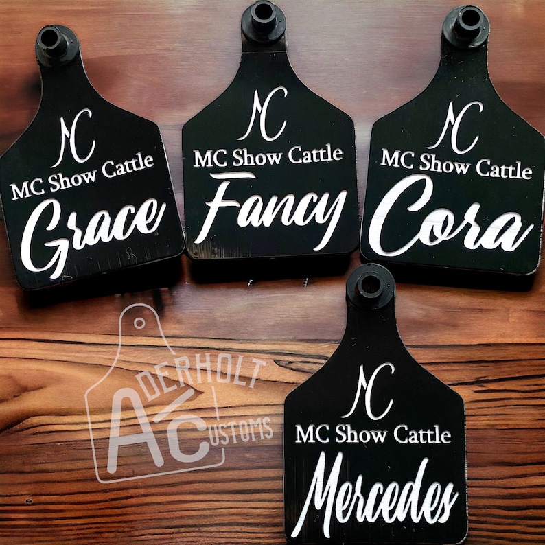 May include: Four black plastic livestock ear tags with a rounded shield shape and a top mounting post. Each tag features the white printed text MC Show Cattle, a logo, and individual names: Grace, Fancy, Cora, and Mercedes. A watermark reads Derholt Customs.