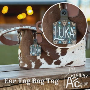 May include: A cowhide bag with a decorative ear tag that says "LUKA". The tag has a pattern of cacti, stars, and longhorn cattle. The bag tag is attached to a brown leather handle. The words "Ear Tag Bag Tag" are at the bottom.