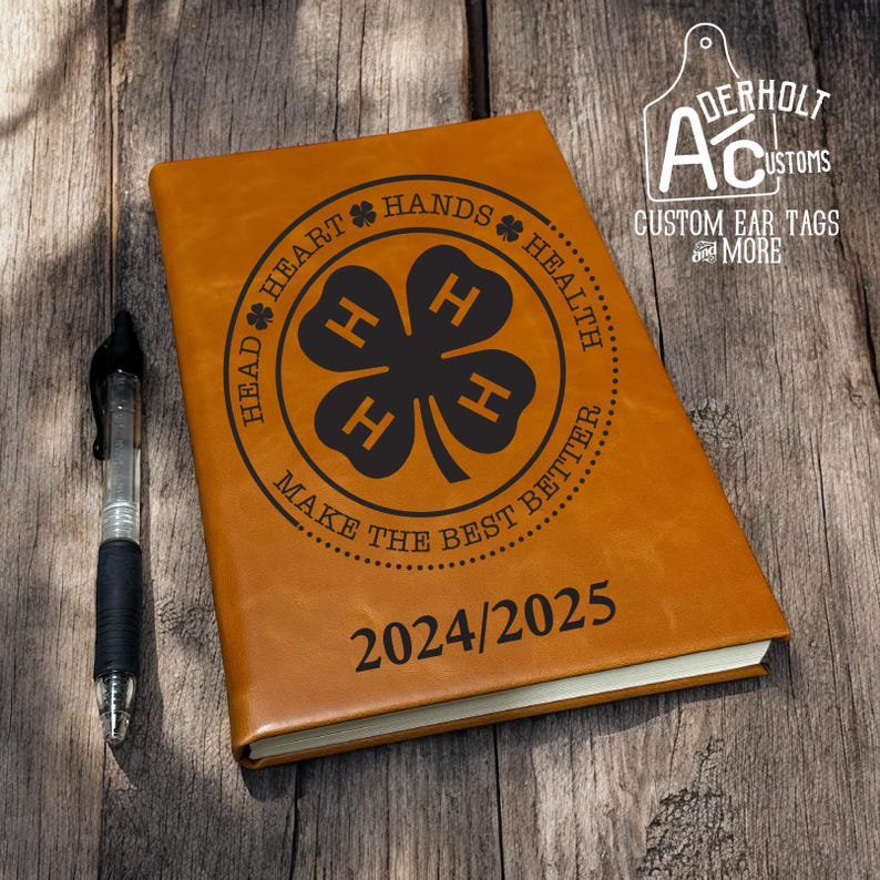 May include: A tan leather-like journal featuring a black circular emblem with a four-leaf clover containing four H letters. The text reads HEAD, HEART, HANDS, HEALTH, MAKE THE BEST BETTER, and 2024/2025. A black pen is positioned to the left of the journal.