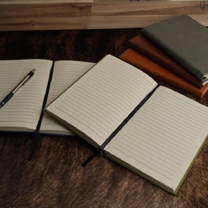 May include: A collection of notebooks with cream-colored lined pages and black ribbon bookmarks. One notebook lies open with a silver and black ballpoint pen resting on the page. A stack of three closed notebooks in gray, dark brown, and tan.