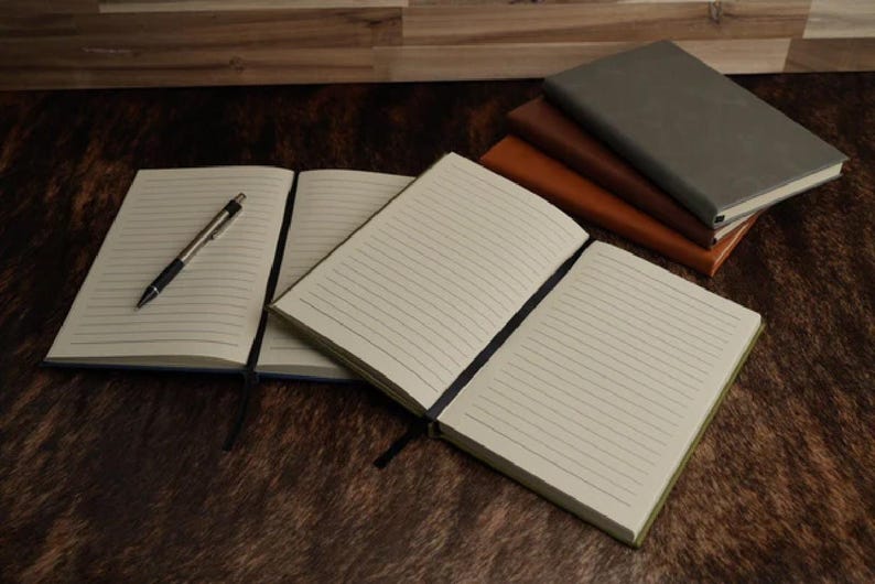 May include: A collection of notebooks with cream-colored lined pages and black ribbon bookmarks. One notebook lies open with a silver and black ballpoint pen resting on the page. A stack of three closed notebooks in gray, dark brown, and tan.