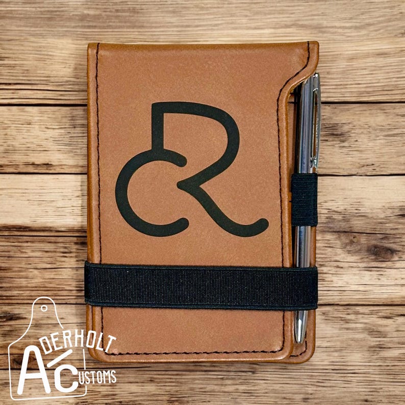 Puede incluir: Una libreta de cuero marr&oacute;n con una banda el&aacute;stica negra y un portaplumas. La libreta tiene un monograma negro de las letras "CR" en la parte delantera. La libreta est&aacute; sobre una superficie de madera.
