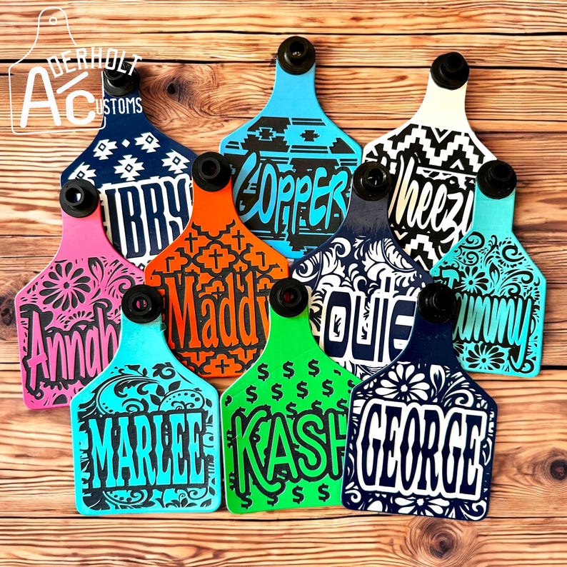 May include: A collection of ten colorful livestock ear tags featuring various patterns and custom names in bold lettering. Each tag has a black circular attachment point at the top, displaying names like Libby, Copper, Wheezy, Annah, Maddy, Outie, Tunny, Marlee, Kash, and George.