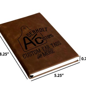 May include: A brown notebook with a black logo featuring the text Aderholt Customs and Custom Ear Tags and More. The notebook measures 8.25 inches in length, 5.25 inches in width, and 0.75 inches in thickness as indicated by the labels.