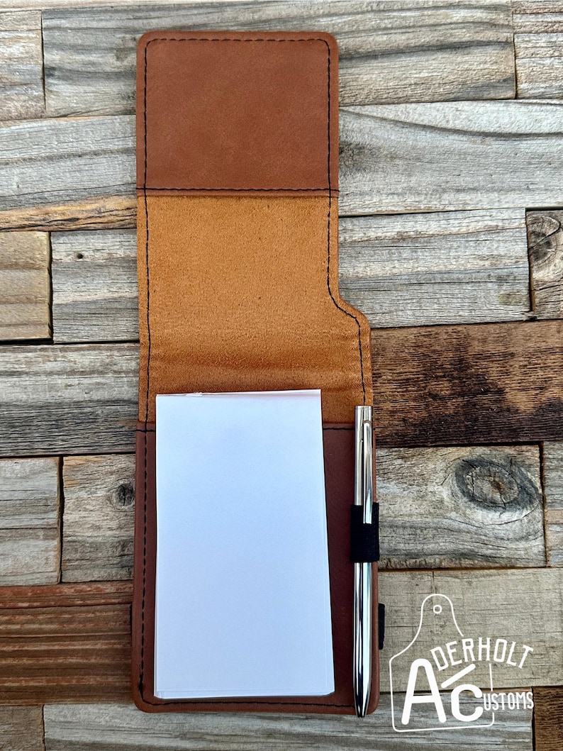 Puede incluir: Una cubierta de cuaderno de cuero marr&oacute;n con un portaplumas y un bloc de notas en blanco en el interior. La cubierta tiene un dise&ntilde;o cosido y una solapa que se pliega sobre el bloc de notas. La marca "ADERHOLT CUSTOMS" es visible en la esquina inferior derecha.