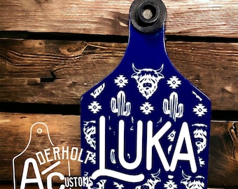 May include: A blue cow tag with white graphics of cacti, longhorn cattle, and geometric patterns. The name "LUKA" is printed in large white letters. A black plastic piece is at the top for hanging.