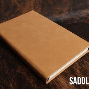 May include: A rectangular notebook with a smooth, solid saddle tan cover and white pages visible along the edge. The front cover is plain and features no additional markings, patterns, or textures. The text SADDLE TAN is printed in white.