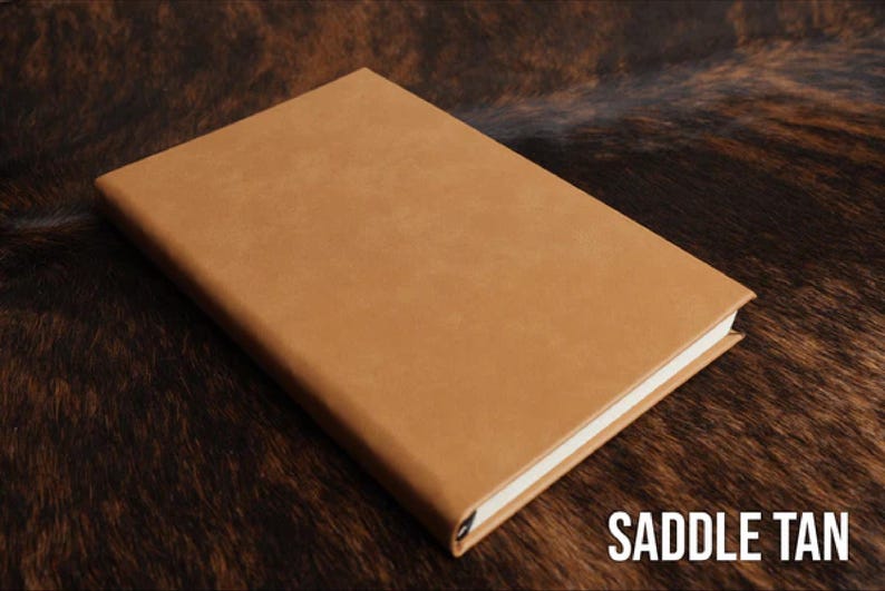 May include: A rectangular notebook with a smooth, solid saddle tan cover and white pages visible along the edge. The front cover is plain and features no additional markings, patterns, or textures. The text SADDLE TAN is printed in white.