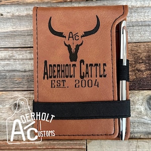 May include: A brown leather notebook with a black elastic band and a silver pen. The notebook is engraved with a black silhouette of a bull skull with horns and the text "Ać", "ADERHOLT CATTLE", and "EST. 2004".