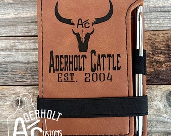 May include: A tan leather notebook cover featuring a laser-engraved bull skull logo with the text Aderholt Cattle Est. 2004. A black elastic band secures the cover, and a silver metal pen is held in a side loop. A logo with the text Aderholt Customs is visible.