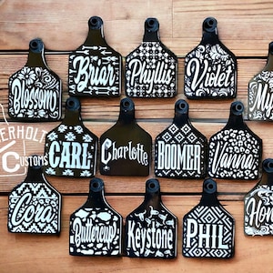 May include: A collection of fourteen black livestock ear tags featuring white custom names and various patterns including floral, geometric, and animal prints. Each tag has a circular hole at the top. The text Aderholt Customs is visible.
