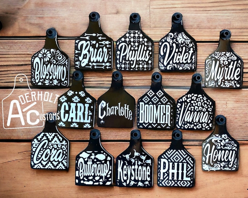 May include: A collection of fourteen black livestock ear tags featuring white custom names and various patterns including floral, geometric, and animal prints. Each tag has a circular hole at the top. The text Aderholt Customs is visible.