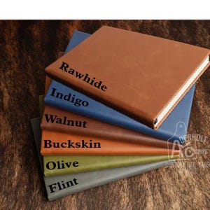 May include: A stack of six rectangular journals with smooth covers in various colors, each labeled with black text: Rawhide, Indigo, Walnut, Buckskin, Olive, and Flint. The journals are arranged in a cascading pile, showing their spines and front covers.