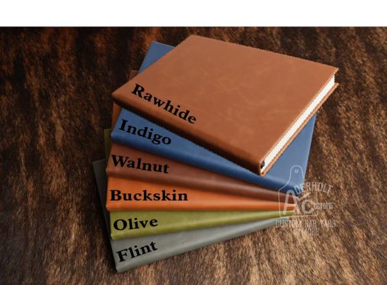 May include: A stack of six rectangular journals with smooth covers in various colors, each labeled with black text: Rawhide, Indigo, Walnut, Buckskin, Olive, and Flint. The journals are arranged in a cascading pile, showing their spines and front covers.