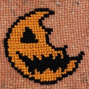 Könnte beinhalten: Eine Nahaufnahme eines Kreuzstich-Designs eines Halbmondes mit einem gruseligen Gesicht. Der Mond ist orange und das Gesicht ist schwarz.