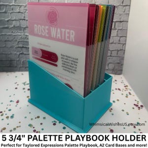 May include: A turquoise palette playbook holder containing a stack of colorful cards. The top card is pink with the words "ROSE WATER" and a rose graphic. The holder is labeled "5 3/4" PALETTE PLAYBOOK HOLDER".
